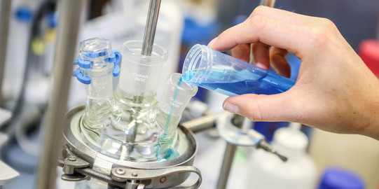 A hand fills a turquoise blue liquid into another glass vessel in the laboratory.