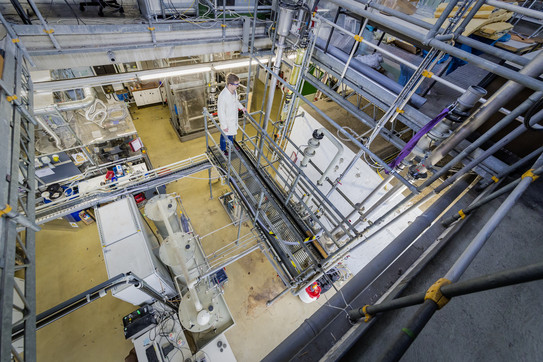 A person with a white coat stands in a laboratory with several large containers and metal pipes.