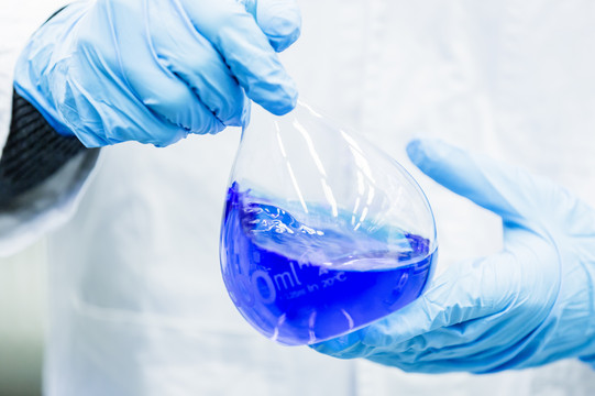 Two hands in blue rubber gloves hold a volumetric flask filled with blue liquid.